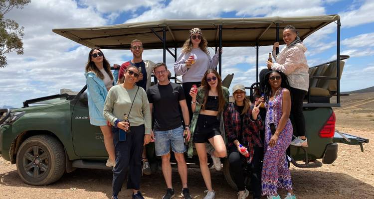 Un groupe de jeunes voyageurs pose devant un véhicule de safari ouvert garé sur une plaine sèche et poussiéreuse sous un ciel partiellement nuageux.