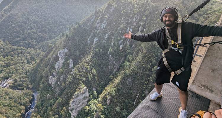 Un homme en harnais se tient sur une haute plateforme de saut à l'élastique, étendant ses bras vers une vaste gorge boisée.
