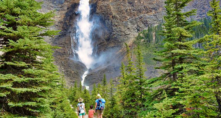 Family walking towards a tall waterfall surrounded by lush trees in Yoho National Park.