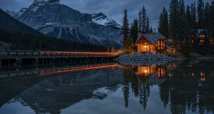 Night view of a cozy cabin and bridge reflecting in a lake, surrounded by mountains.