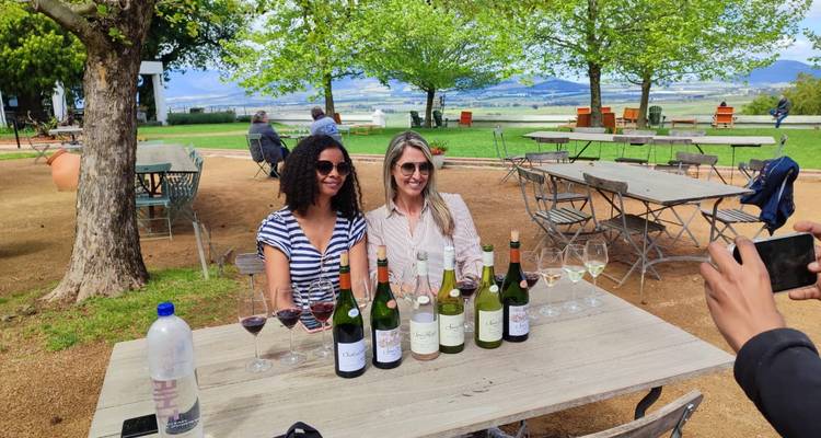 Deux femmes dégustent du vin en plein air dans un domaine viticole pittoresque, avec des bouteilles et des verres alignés sur la table en bois.