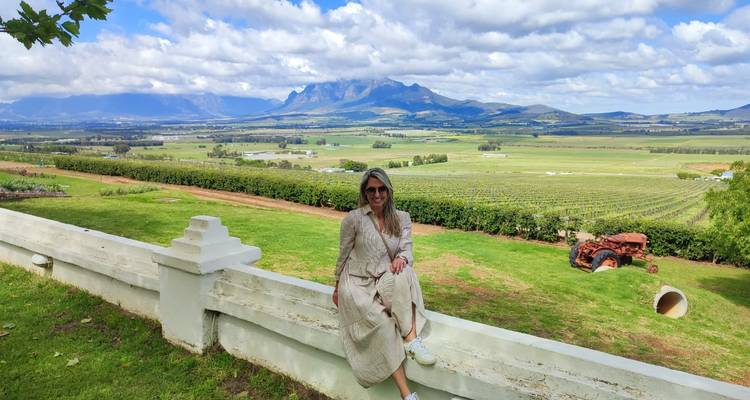 Une femme vêtue d'une robe fluide est assise sur un mur blanc bas surplombant des vignobles ondulants et des montagnes lointaines sous un ciel partiellement nuageux.