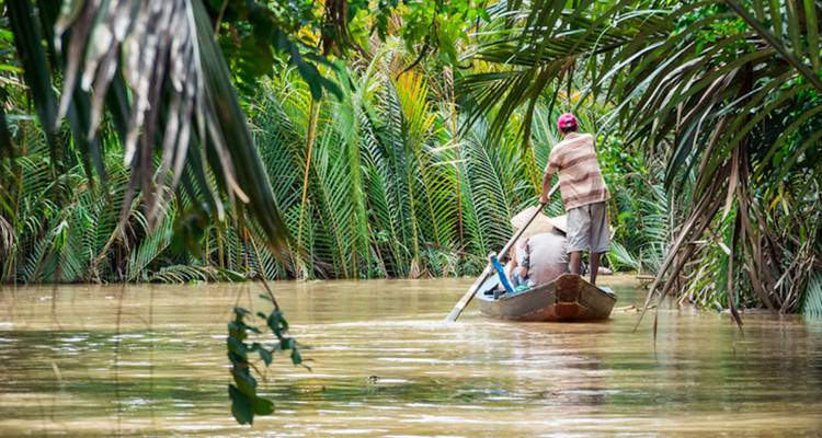 Menschen navigieren einen Fluss in einem kleinen Holzboot, umgeben von üppigem Laubwerk.