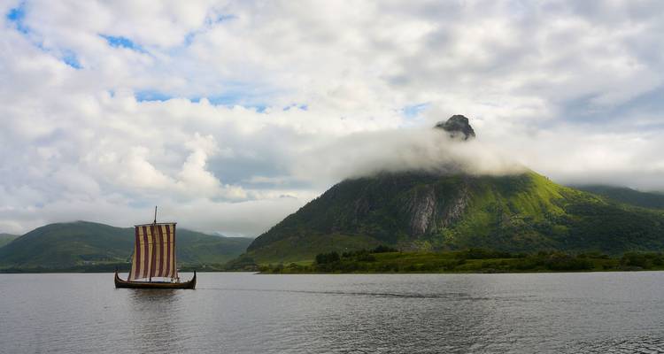 Ein traditionelles Wikingerschiff segelt über einen nebelverhangenen Fjord mit einem steilen grünen Berg, der teilweise von Wolken verhüllt ist.
