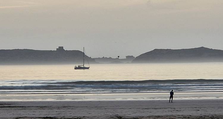 Une scène côtière avec une personne sur la plage et un voilier au loin.