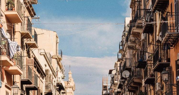 Smalle Mediterrane straat omzoomd door hoge appartementengebouwen met gietijzeren balkons onder een heldere blauwe lucht.