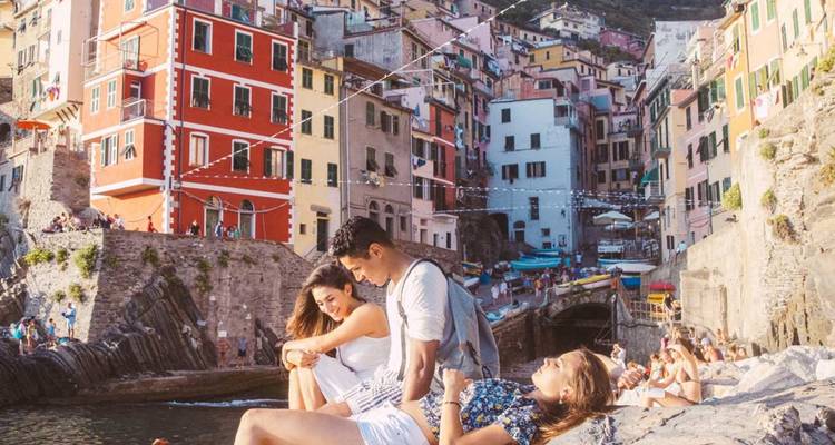 Travellers relax on rocky harbour walls beneath the tall pastel houses of Riomaggiore in Cinque Terre