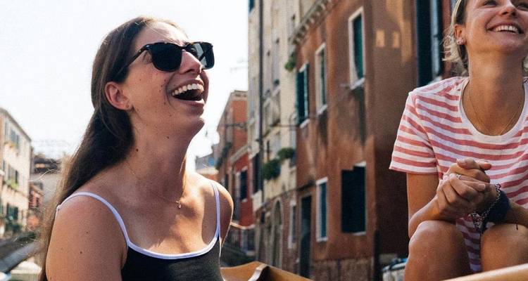 Dos mujeres sonrientes disfrutan de un paseo en bote por un estrecho canal veneciano rodeado de edificios coloridos