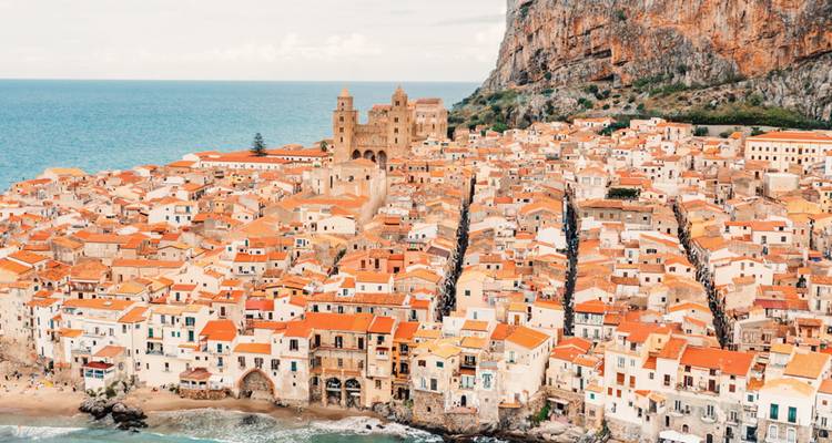 Panoramic view of Cefalù’s orange-tiled rooftops clustered beneath a towering seaside cliff and medieval cathedral