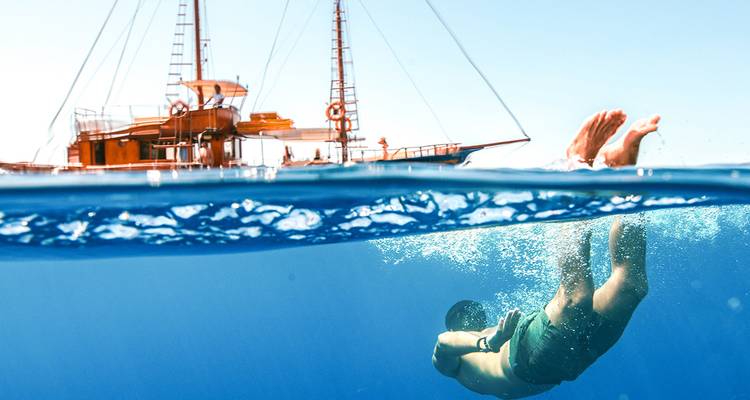 Foto de nivel dividido de un nadador buceando bajo el mar azul cristalino junto a un velero clásico de madera.