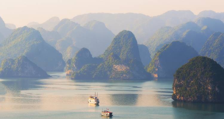 Lumière matinale voilée au-dessus d'une baie tranquille avec des montagnes calcaires en strates et deux navires de croisière.
