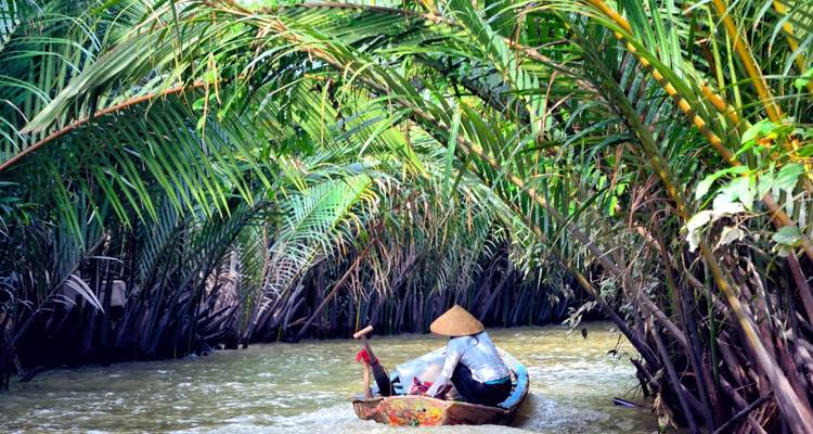 Personne portant un chapeau conique pagayant dans une petite embarcation à travers un canal luxuriant bordé de palmiers.