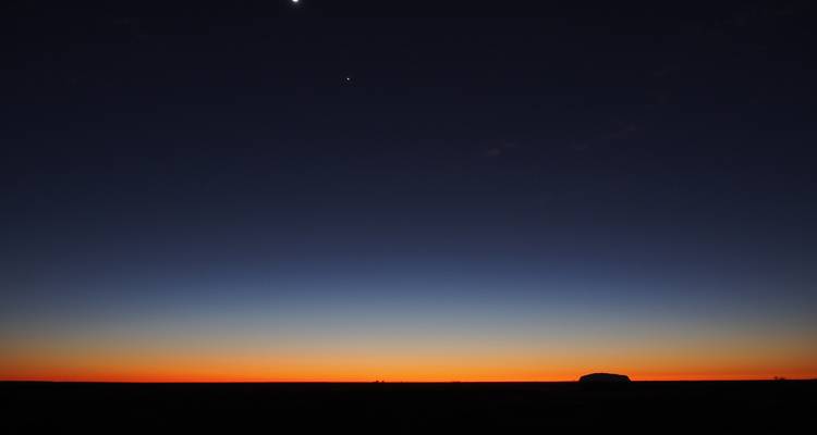 Scène d'aube minimaliste avec la silhouette d'Uluru à l'horizon sous un ciel en dégradé profond passant de l'orange au bleu foncé.