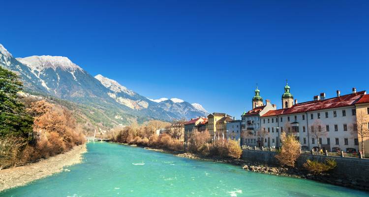 Rivière de montagne coulant près de bâtiments historiques avec des sommets enneigés en arrière-plan.