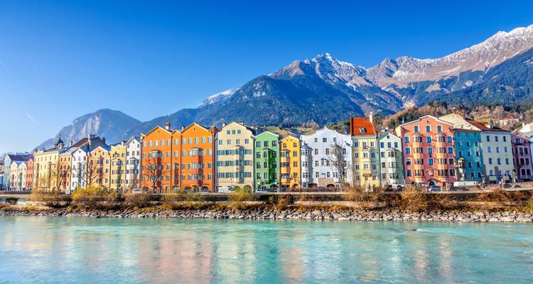 Rangée colorée de bâtiments au bord d'une rivière, avec des montagnes en arrière-plan.