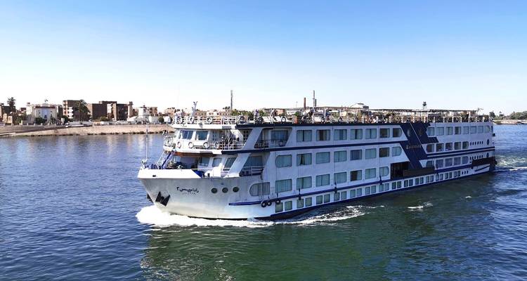 A large cruise ship sailing on a river with buildings on the riverbank.