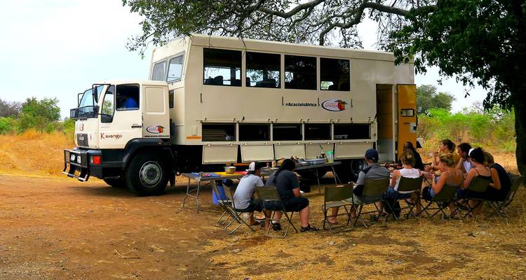 Reizigers zittend op opvouwbare stoelen naast een overland truck in een droog veld