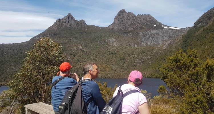 Drei Personen sitzen auf einer Bank mit Blick auf Berge und einen See.