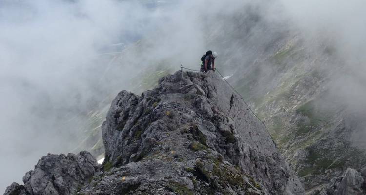 Ein Bergsteiger bewältigt einen schmalen felsigen Grat, der von wirbelndem Bergnebel umhüllt ist.
