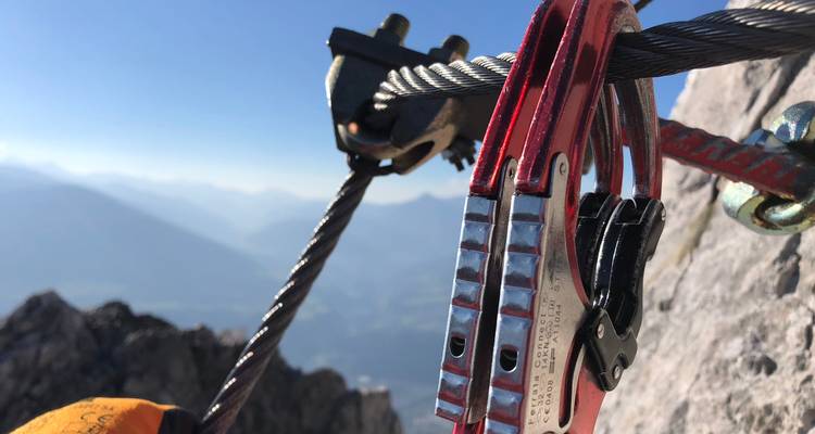 Nahaufnahme einer Klettersteig-Umlenkrolle, die an einem Stahlseil befestigt ist, mit verschwommenen alpinen Ausblicken im Hintergrund.