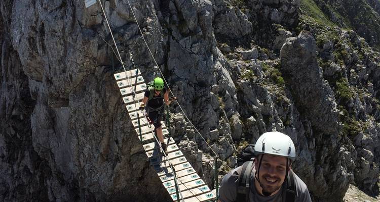 Helmtragende Kletterer überqueren eine schmale Hängebrücke hoch an einer Felswand.