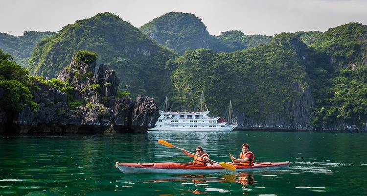Zwei Personen beim Kajakfahren mit einem Kreuzfahrtschiff im Hintergrund in der Halong-Bucht.
