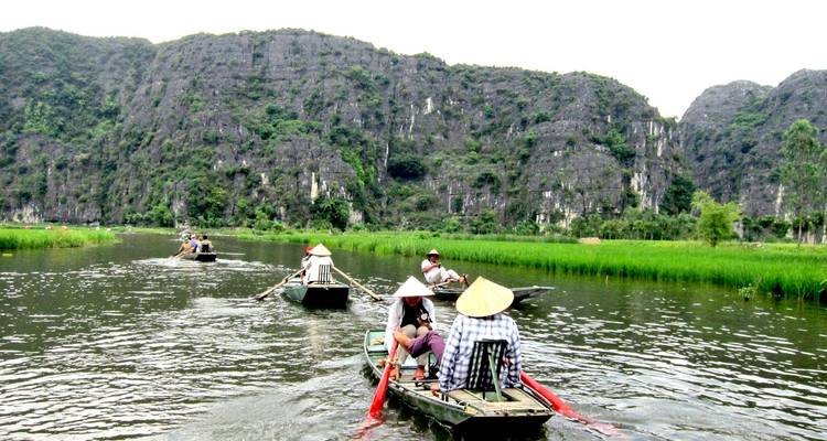 People rowing boats along a river with mountains in the background.