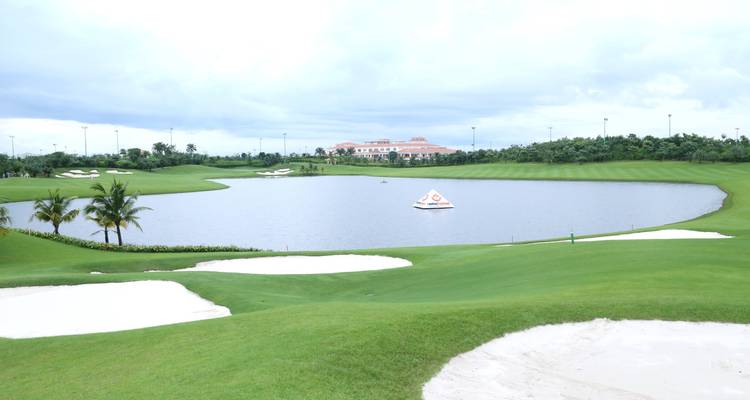 Golfplatz mit einem Teich, Clubhaus und Sandbunkern unter einem bewölkten Himmel.