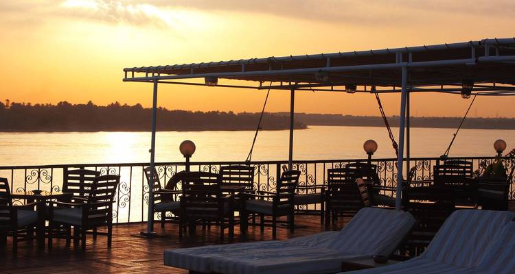 Vue de coucher de soleil sur une rivière calme avec des chaises longues sur une terrasse.
