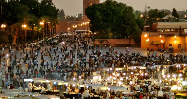 Une vue panoramique nocturne d'une grande place bondée de monde et d'étals de nourriture illuminés, avec une haute tour et des palmiers en arrière-plan.