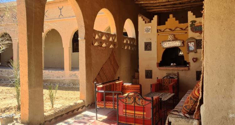 Une terrasse rustique ombragée avec des coussins colorés et des arches donnant sur une cour désertique ; filigrane visible.