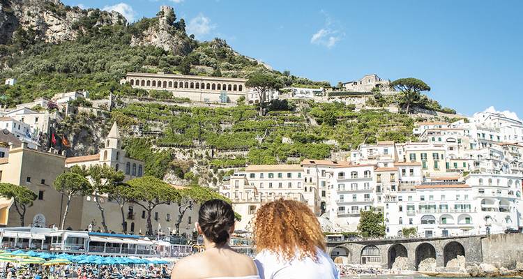 Dos mujeres se relajan en una playa mirando hacia los pueblos en terrazas de las laderas de la Costa Amalfitana.