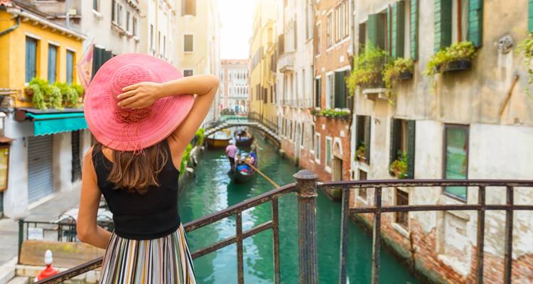 Un voyageur avec un chapeau rose vif se tient sur un pont surplombant un étroit canal vénitien avec des gondoles en contrebas.