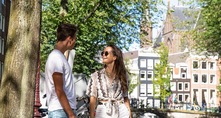 Smiling couple chatting beside a leafy canal with classic gabled houses in Amsterdam.