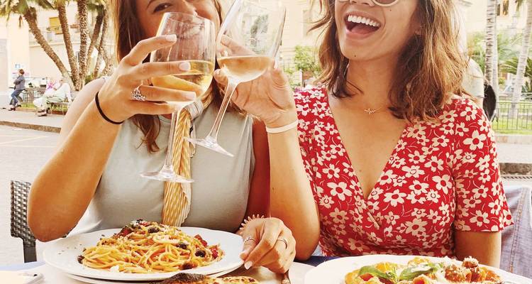 Deux femmes trinquant avec du vin blanc au-dessus d'assiettes de pâtes fraîches dans une trattoria italienne en plein air.