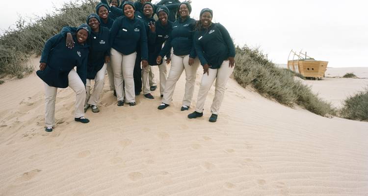 Gruppe von Menschen, die auf Sanddünen stehen mit einem Schiffswrack im Hintergrund.