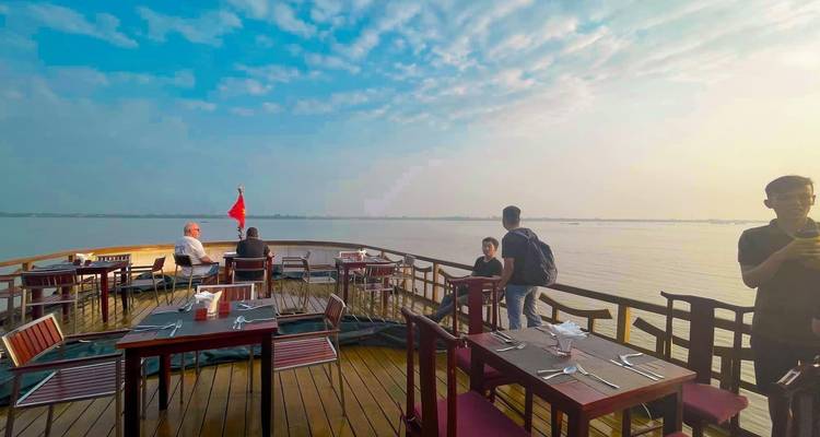 Pont de bateau avec des tables, des chaises, et des personnes profitant d'une vue panoramique sur la rivière avec un drapeau.