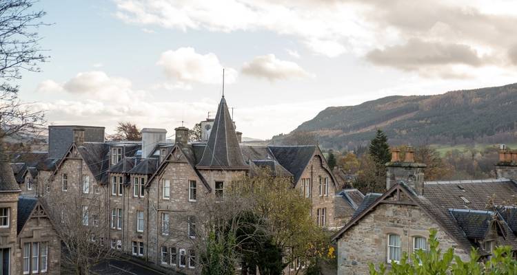 De charmants bâtiments en pierre et toits d'une ville des Highlands se dressent devant des collines boisées et un ciel nuageux.