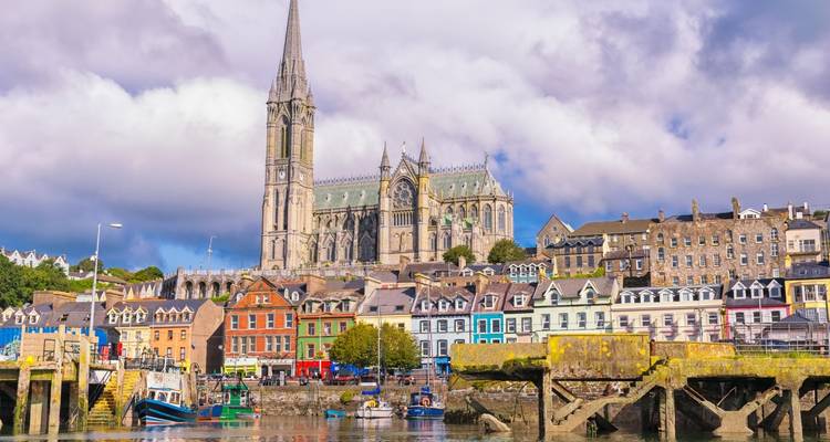 Ville colorée au bord de l'eau couronnée par une imposante cathédrale néo-gothique sous des nuages dramatiques.
