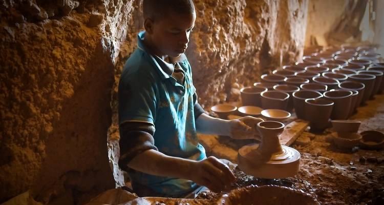 Person crafting pottery indoors with rows of clay pots.