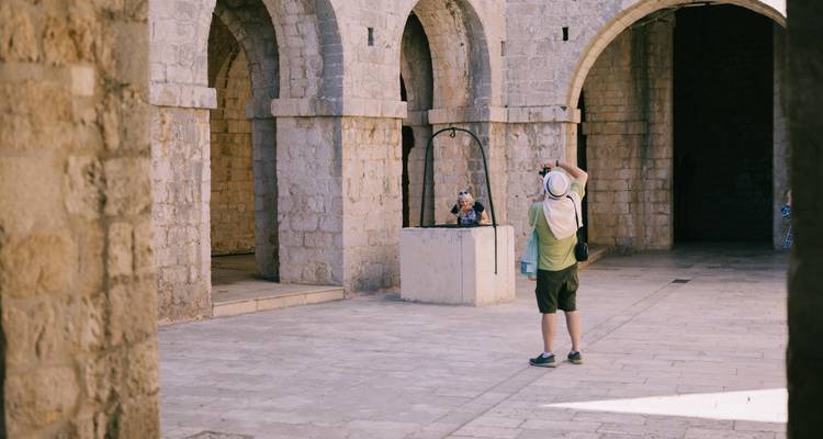 Un voyageur photographie un compagnon en pose à côté d'un ancien puits en pierre dans une cour voûtée.
