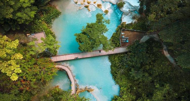 Vue aérienne de bassins de calcaire turquoise reliés par une passerelle en bois au milieu d'une forêt verdoyante.