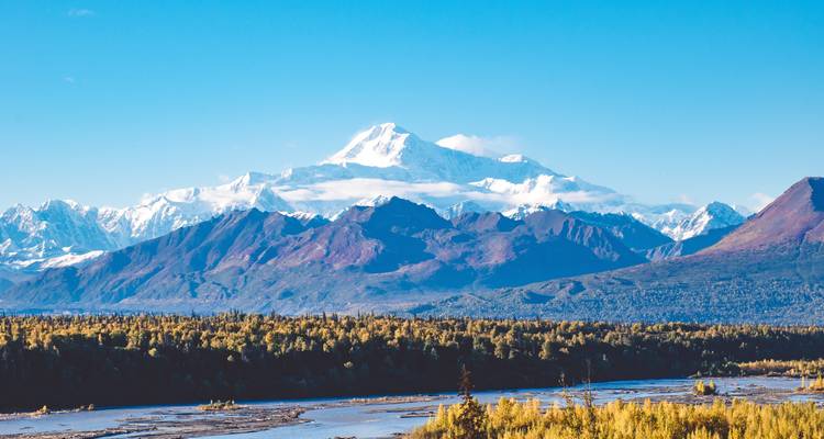 Snow-capped Denali dominates the horizon above forested foothills and a broad braided river.