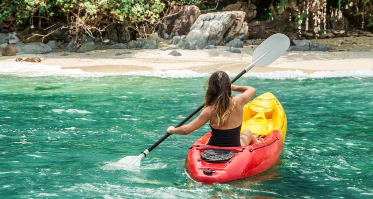 Mujer remando en un kayak rojo sobre agua turquesa cristalina hacia una playa tropical de arena.