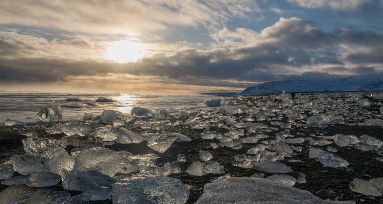 Eisstücke auf einem schwarzen Sandstrand bei Sonnenuntergang.
