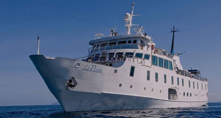 Embarcación de expedición blanca 'La Pinta' anclada en el océano azul profundo bajo un cielo ecuatorial despejado.