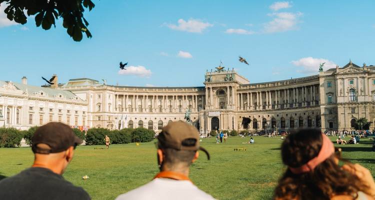 Des touristes assis sur une pelouse devant un impressionnant complexe de bâtiments.