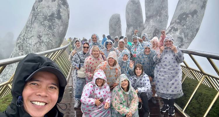 Grupo de turistas sonrientes con ponchos de lluvia coloridos posando en el brumoso Puente Dorado sostenido por manos gigantes de piedra.