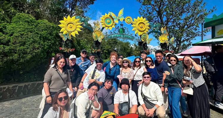 Grupo de turistas posando bajo un arco decorativo de girasoles en un día de cielo azul brillante.