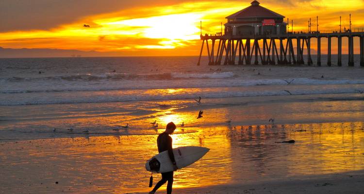 Surfeur marchant sur une plage au coucher du soleil avec une jetée en arrière-plan.